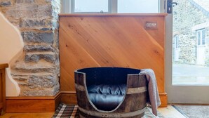 A rustic room with a stone wall, a wooden partition, a barrel-shaped dog bed, and a metal bowl on a plaid floor. - 1 Bed in Launceston (oc-81332) (Saint Giles on the Heath)