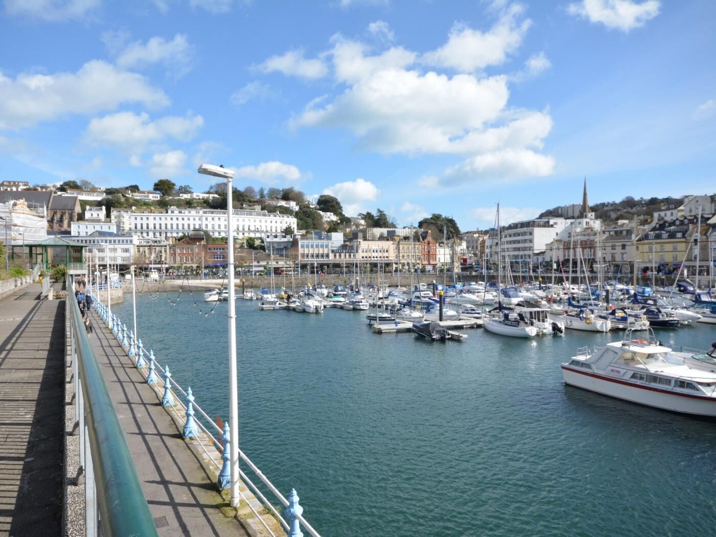 A marina with boats docked, a promenade with railings, and buildings in the background.