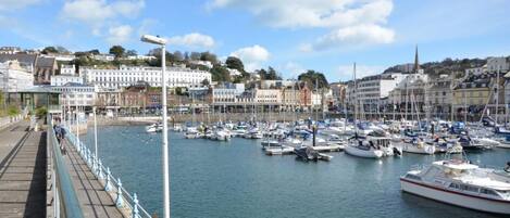 A marina with boats docked, a promenade with railings, and buildings in the background.