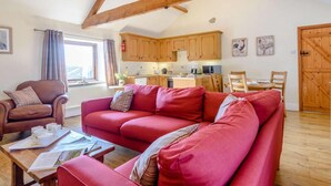 Ein gemütliches Wohnzimmer mit einem roten Sofa, einem braunen Ledersessel und einem Holzesstisch. Im Hintergrund ist eine Küche mit Holzschränken und einem Fenster zu sehen.