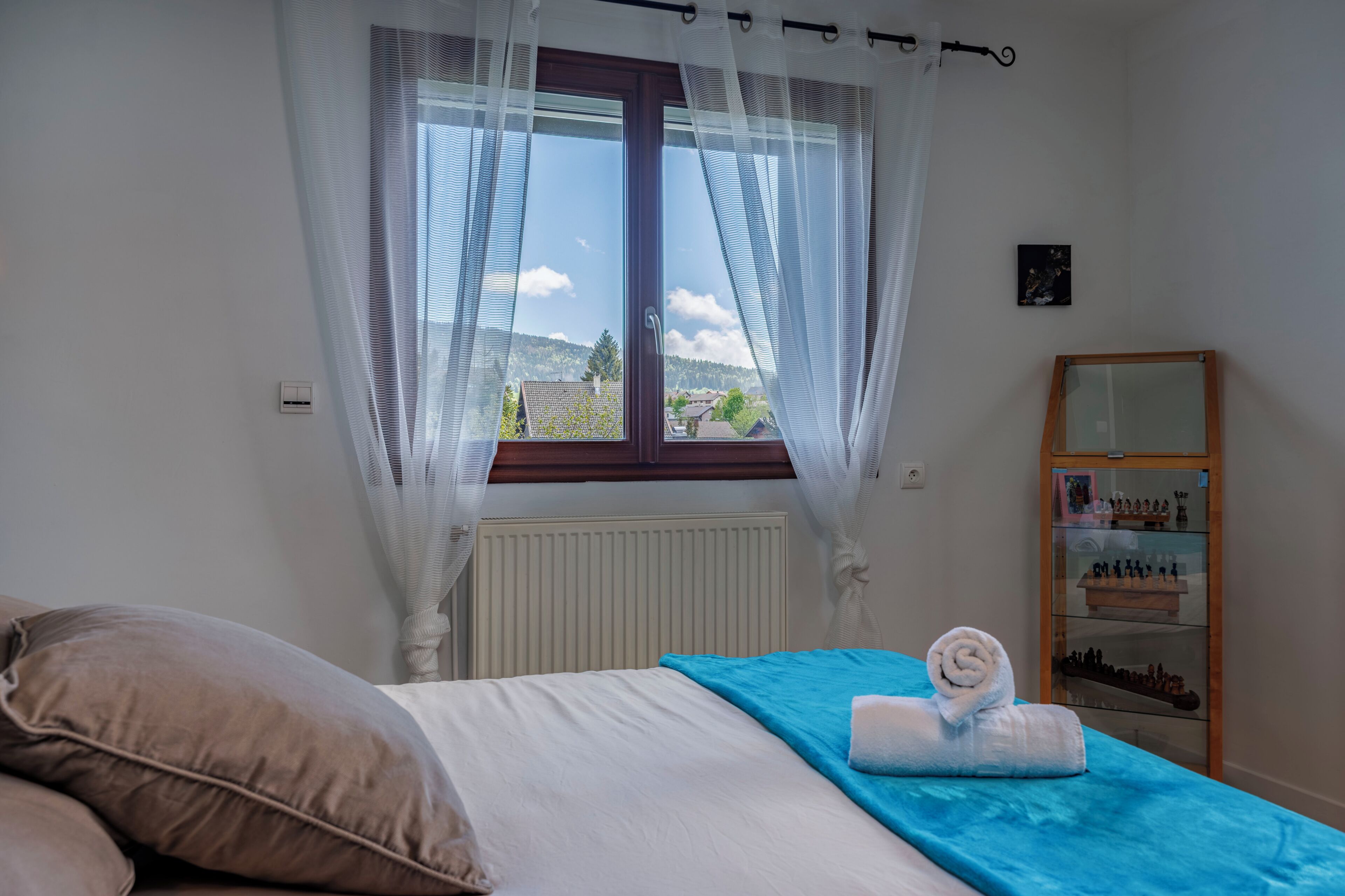 A bedroom with a bed, a window with sheer curtains, a radiator, a wooden shelf with chess pieces, and a small framed picture on the wall.