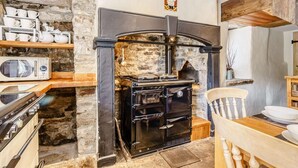A rustic kitchen with a stone wall, a black stove, and a wooden table with chairs. - 5 Bed in Richmond (oc-81457) (Gunnerside)