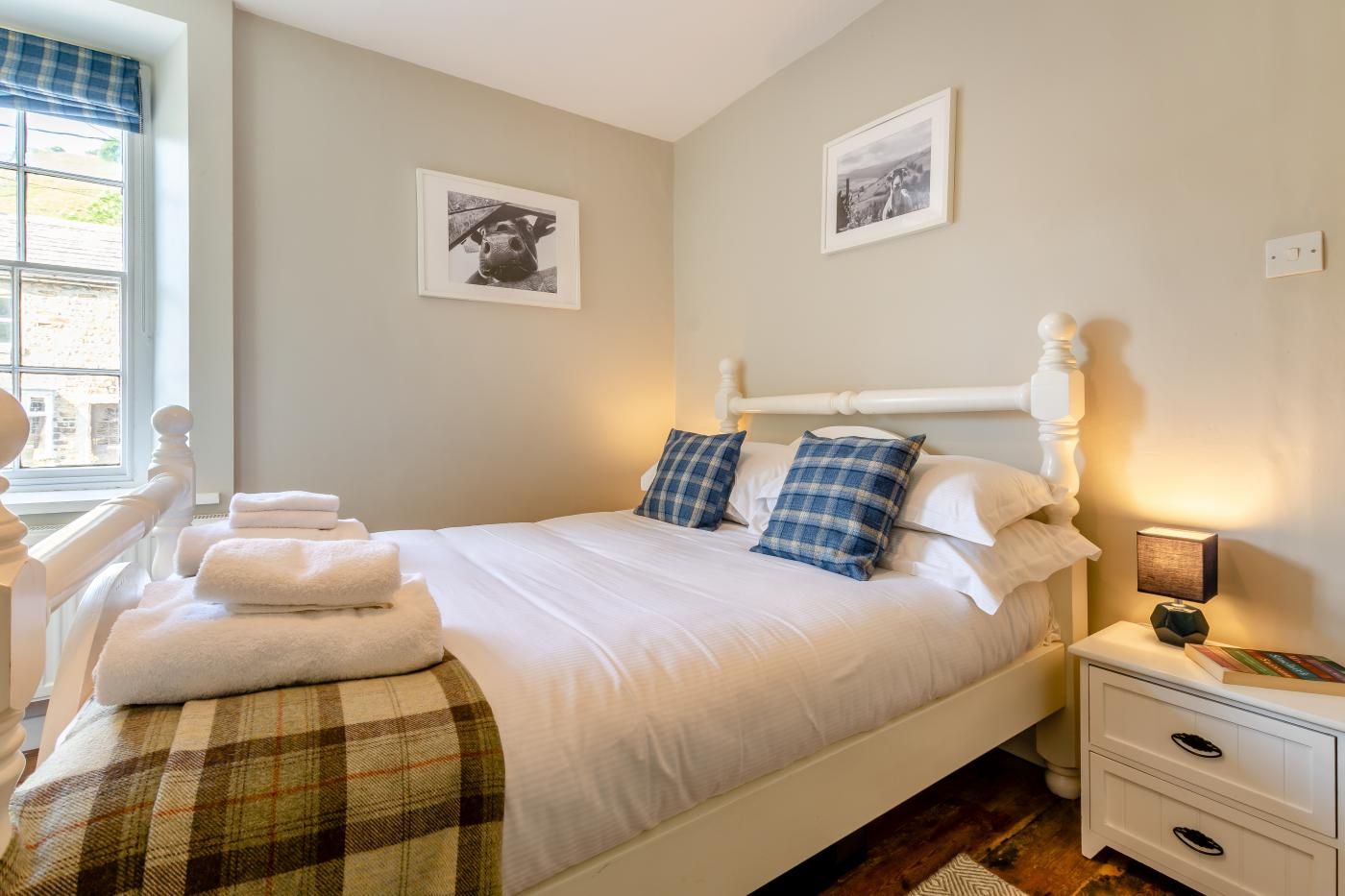 Een net opgemaakt bed met witte lakens en blauw geruit kussens, een nachtkastje met een lamp, en twee ingelijste foto's aan de muur.