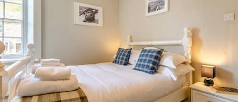 Een net opgemaakt bed met witte lakens en blauw geruit kussens, een nachtkastje met een lamp, en twee ingelijste foto's aan de muur.