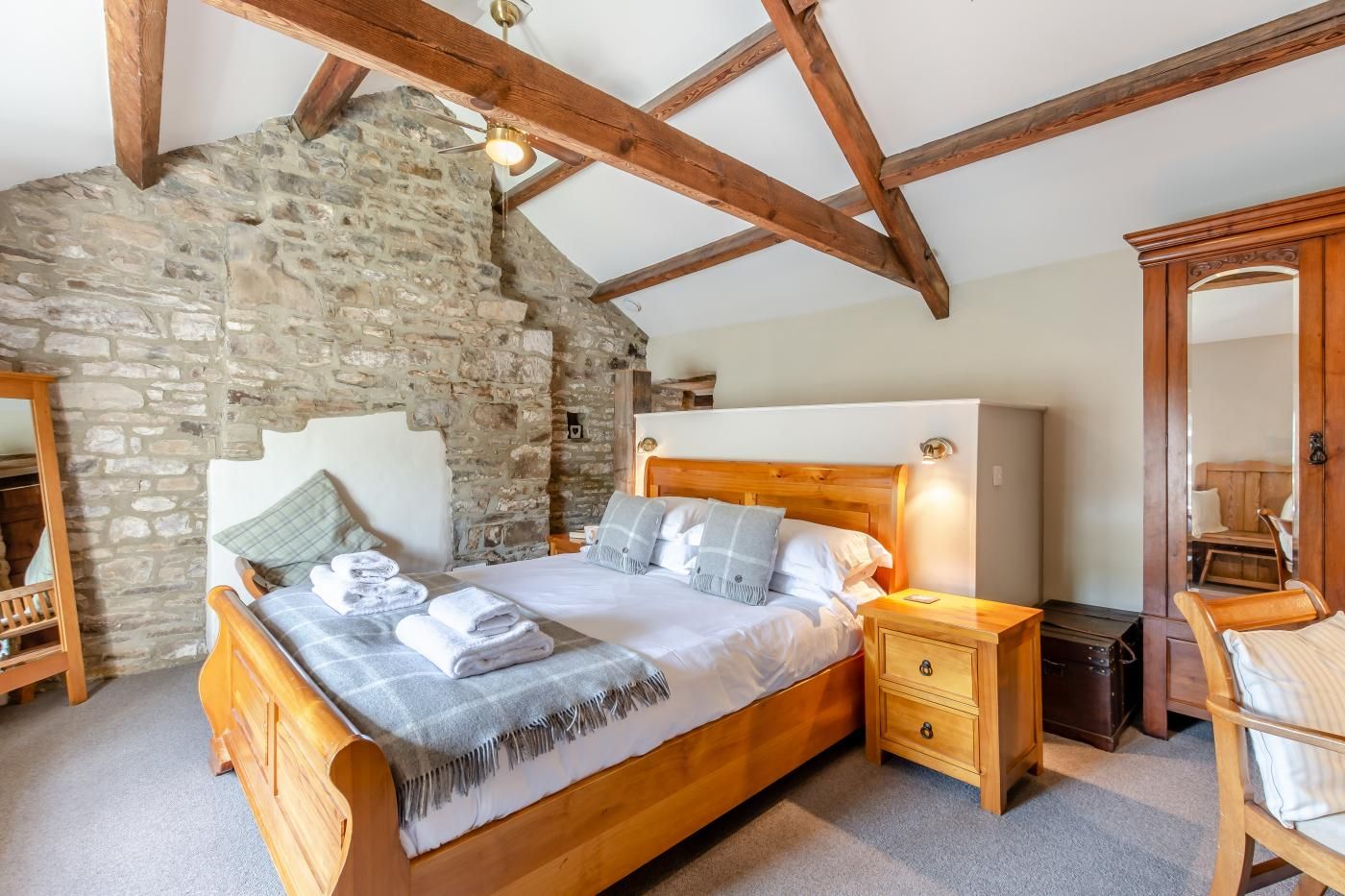Een slaapkamer met een stenen muur, houten balken, een bed met kussens, een nachtkastje, een houten stoel en een spiegel.