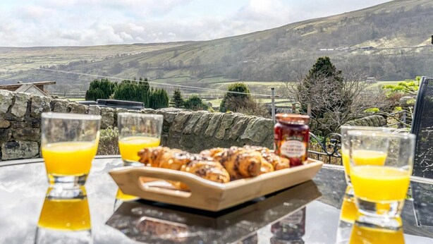 A table with a tray of pastries, glasses of orange juice, and a jar of jam, overlooking a scenic landscape. - 5 Bed in Richmond (oc-81457) (Gunnerside)