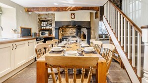 A dining area with a wooden table and chairs, a stone fireplace, and a kitchen in the background. - 5 Bed in Richmond (oc-81457) (Gunnerside)