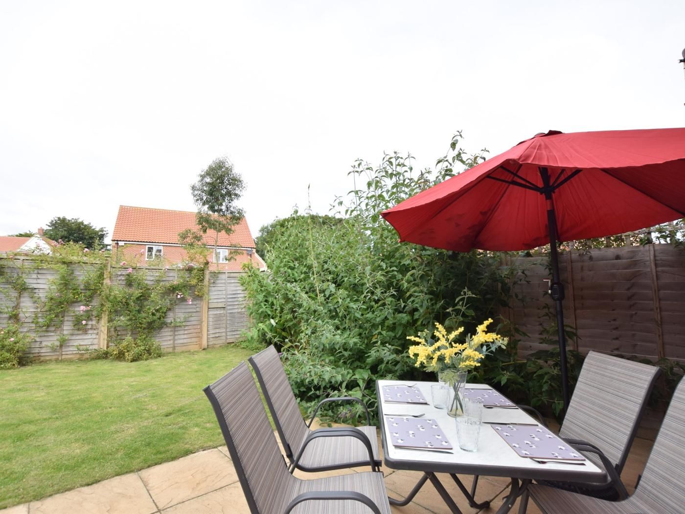 En udendørs terrasse med et bord, stole, en rød parasol og en vase med gule blomster.