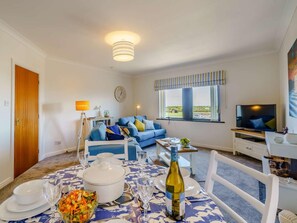 A living room with a sofa, a dining table set with plates, glasses, and a bottle, and a television on a stand. - 2 Bed in Maryport (oc-81443) (Maryport)