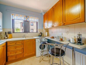 A kitchen with wooden cabinets, a sink, a window with a view, a washing machine, and two bar stools. - 2 Bed in Maryport (oc-81443) (Maryport)