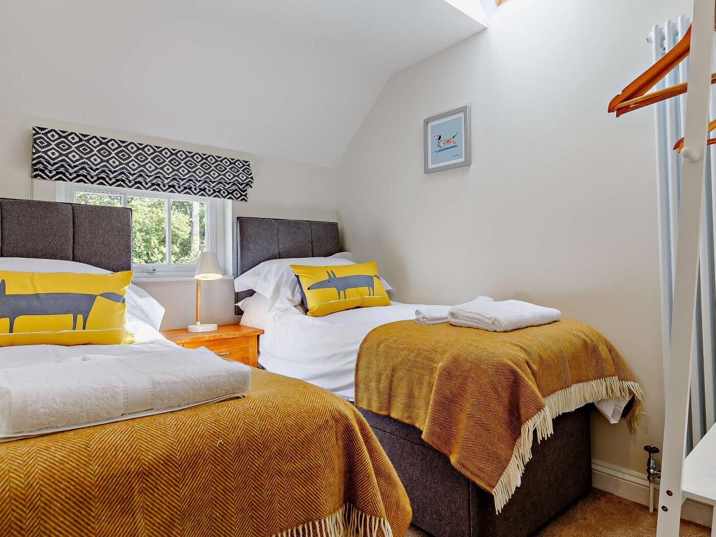 Un dormitorio con dos camas, una ventana con cortinas estampadas, una lámpara de noche y un cuadro enmarcado en la pared.