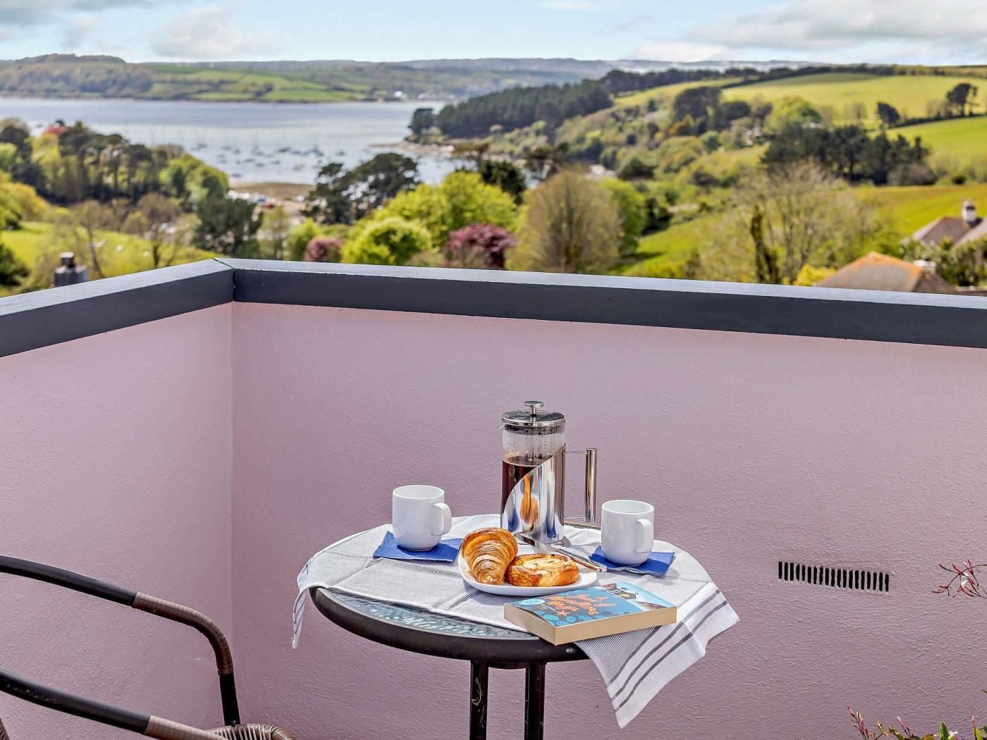 Un balcon avec une table préparée pour le petit-déjeuner, un livre et une vue sur un lac et des collines.