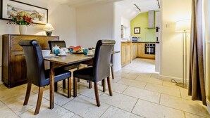 Un comedor con una mesa de madera, sillas y un florero. Se ve una cocina al fondo, con un microondas y armarios.