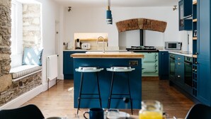 A modern kitchen with a central island, bar stools, a stone wall, and a view of the outdoors. - 8 Bed in Newquay (oc-81510) (Quoit)