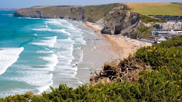 A coastal beach with rocky cliffs, a sandy shore, and a building complex with multiple levels. - 8 Bed in Newquay (oc-81510) (Quoit)