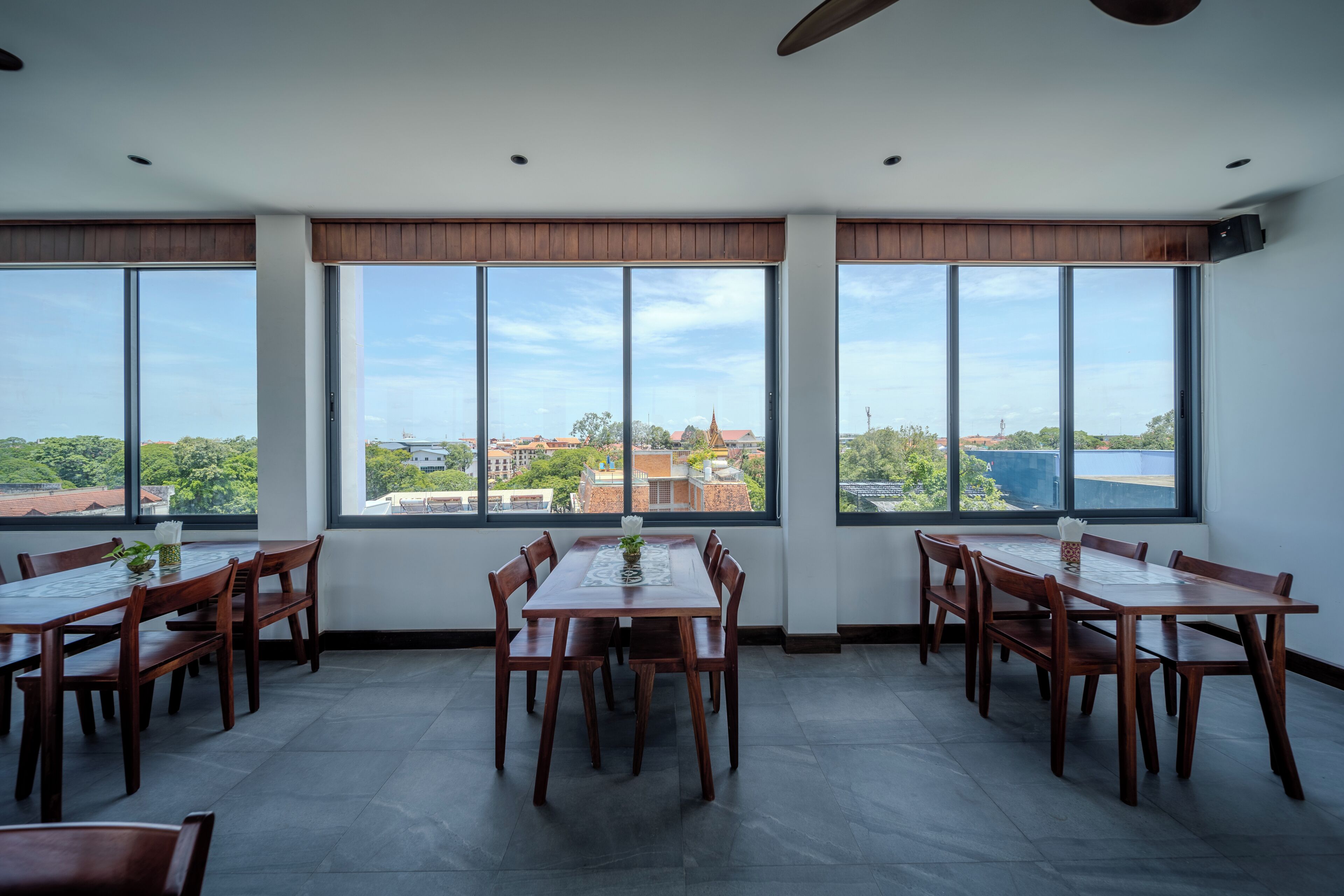 Un comedor con una mesa grande y sillas, ventanas amplias con vistas a edificios y árboles, y un ventilador de techo.