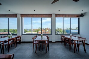 Un comedor con una mesa grande y sillas, ventanas amplias con vistas a edificios y árboles, y un ventilador de techo.
