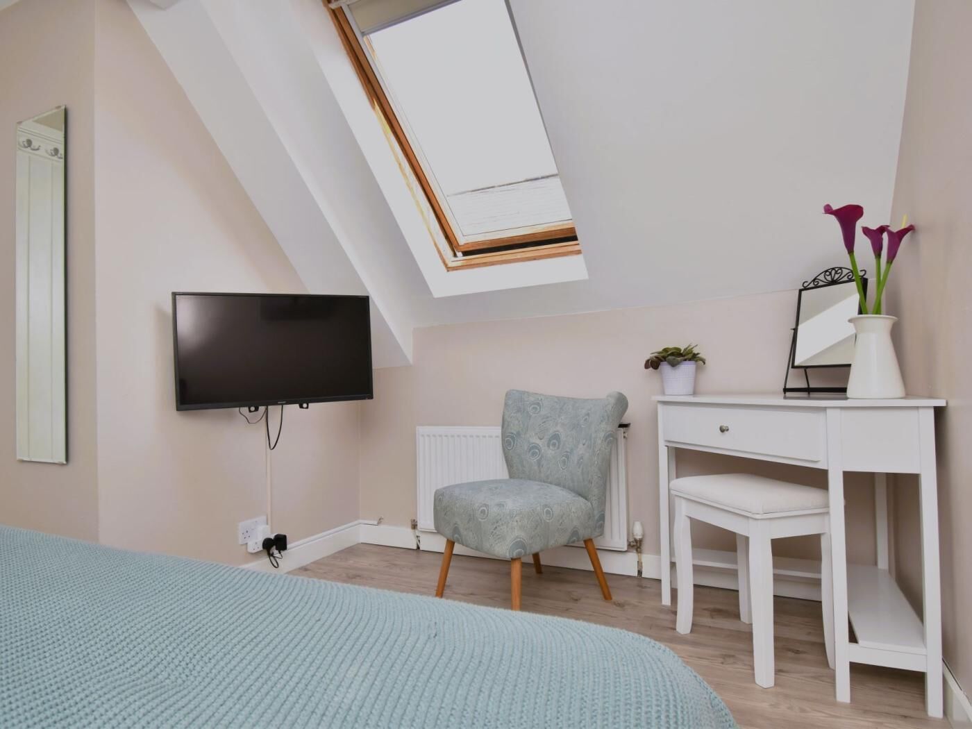 A modern bedroom with a skylight, a flat-screen TV, a grey armchair, a white desk, and a bedside table with a vase of flowers.