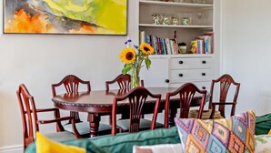 A dining area with a round wooden table, six chairs, a vase with sunflowers, a bookshelf with books and decorative items, and a large abstract painting on the wall.