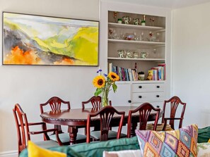 A dining area with a round wooden table, six chairs, a vase with sunflowers, a bookshelf with books and decorative items, and a large abstract painting on the wall.