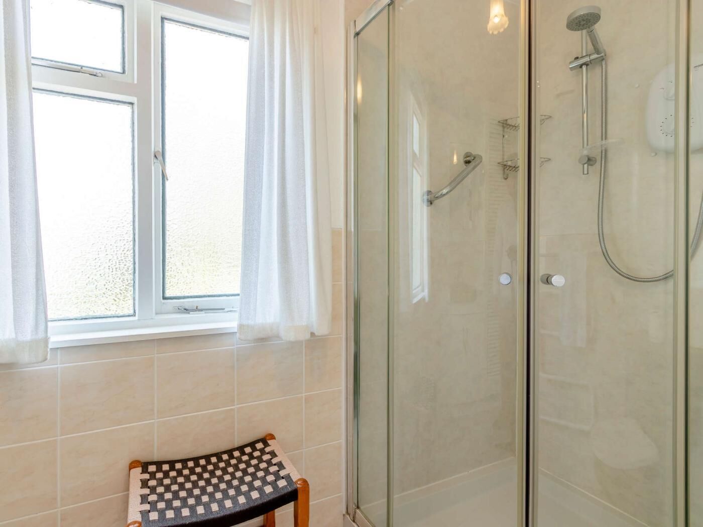 Ducha con cerramiento de vidrio, ducha de mano y grifo de mano, ventana con cortinas blancas y una pequeña silla con asiento a cuadros.