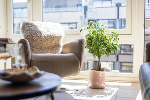 A modern living room with a leather armchair, a potted plant, and a view of the city through large windows. - 1 bedroom accommodation in Oostduinkerke (Oostduinkerke)
