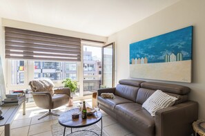 A modern living room with a leather sofa, a coffee table, and a large window with blinds. - 1 bedroom accommodation in Oostduinkerke (Oostduinkerke)