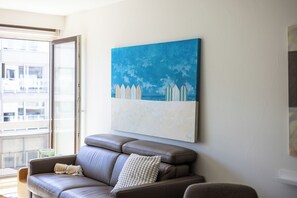 A modern living room with a brown leather sofa, a large abstract painting, and a view of a building through a glass door. - 1 bedroom accommodation in Oostduinkerke (Oostduinkerke)