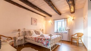 Ein Schlafzimmer mit Holzboden, einem Bett mit floralem Bettzeug, einem Nachttisch mit Lampe, einem Sessel und einem Fenster mit Vorhängen.