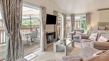 Un salon spacieux avec un canapé, une table basse et un téléviseur à écran plat. Il y a un balcon avec une table et des chaises, et une vue sur la verdure environnante.