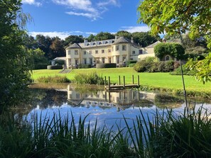 Ein großes, mehrgeschossiges Gebäude mit symmetrischer Fassade, ein Teich im Vordergrund und ein gepflegter Garten.