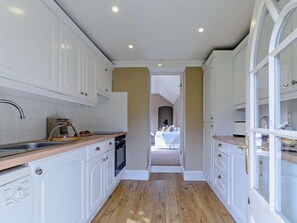 Eine schmale Küche mit weißen Schränken, einer Holz Arbeitsplatte und Blick in ein Schlafzimmer durch eine geöffnete Tür.