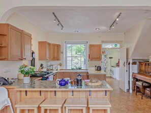 A kitchen with wooden cabinets, a central island with stools, a window, and a view of greenery outside. - 4 Bed in St. Austell (oc-82414) (Coombe)