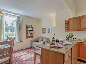 A cozy kitchen with a wooden island, a dining area with a table and chairs, a bookshelf, and a window with curtains. - 4 Bed in St. Austell (oc-82414) (Coombe)