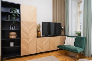 Ein modernes Wohnzimmer mit Fernseher, einem grünen Sofa, einem Holzschrank mit Fischgrätmuster und einem Fenster mit Vorhängen.