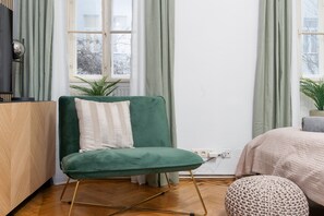 Ein modernes Schlafzimmer mit einem grünen Sessel, einer Holzkommode, einem Nachttisch und einem Fenster mit Vorhängen.