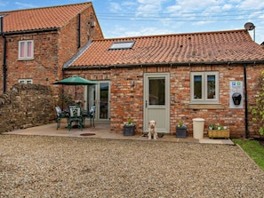 A brick house with a tiled roof, a patio with outdoor furniture, and a dog. - 2 Bed in Northallerton (oc-82411) (Ainderby Steeple)