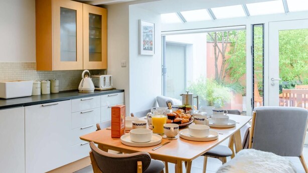 Eine moderne Küche mit weißen Schränken, einem Frühstückstisch und Blick auf einen Garten durch die Glastür.