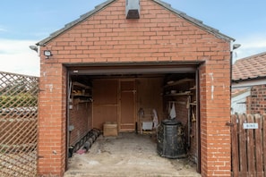 Ein Backsteinhaus mit geöffneter Garagentür, die einen unordentlichen Innenraum mit Regalen, einem Fass und einem Schild mit der Aufschrift 'BINS' zeigt.