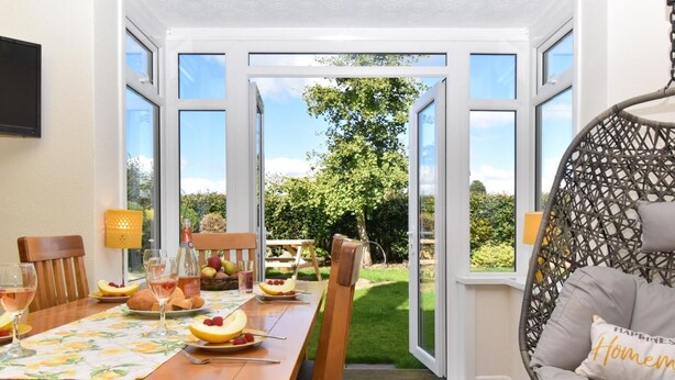 Un coin repas avec une table dressée pour deux personnes, une chaise en osier et une vue sur un jardin, visible à travers de grandes fenêtres.
