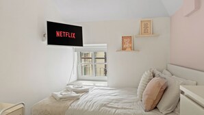 A bedroom with a bed, a TV mounted on the wall displaying 'NETFLIX', a window, and a shelf with decorative items. - Modern Edinburgh City Escape | Sleeps 2 (Edinburgh)