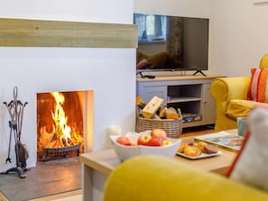A living room with a fireplace, a television on a stand, a yellow sofa, and a table with fruits and pastries. - 2 Bed in Wadebridge (oc-82681) (Little Petherick)