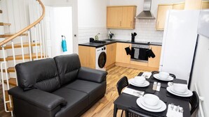 Een compacte leefruimte met een zwarte leren bank, een eettafel voor vier personen, een wasmachine en houten kasten.