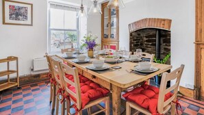 A dining area with a large wooden table set for four, red cushioned chairs, hanging pendant lights, a stone fireplace, and a window with a view of a vehicle outside. - 4 Bed in Cardigan (oc-82766) (Rhos-hill)