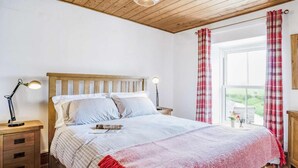 A bedroom with a wooden headboard, a bed with a white and red blanket, a nightstand with a lamp, and a window with curtains. - 4 Bed in Cardigan (oc-82766) (Rhos-hill)