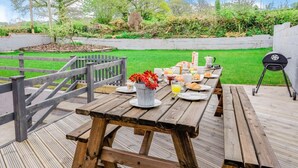 An outdoor dining area with wooden tables and benches, a barbecue grill, and a garden with a wooden fence and greenery. - 4 Bed in Cardigan (oc-82766) (Rhos-hill)