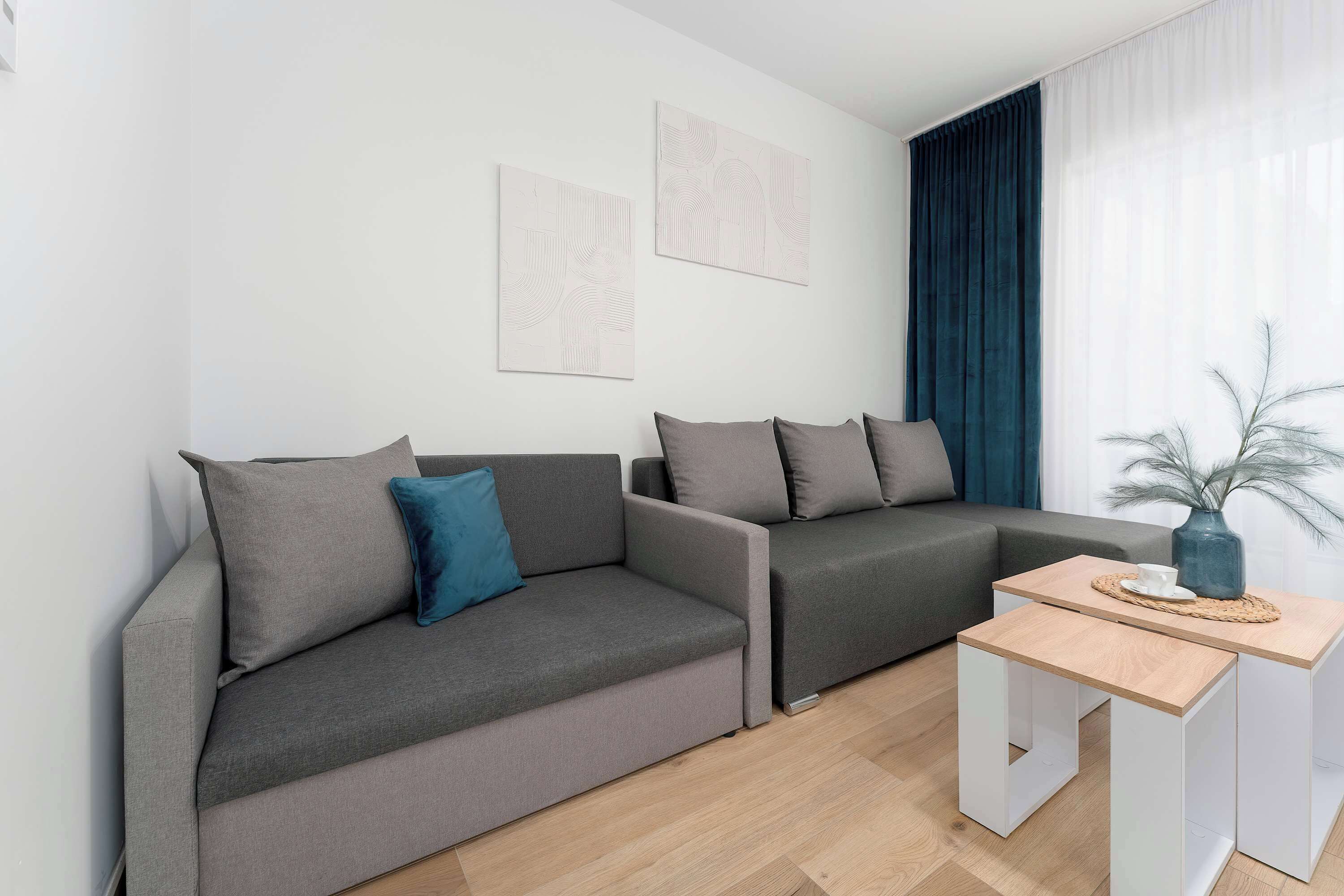 A modern living room features a grey sectional sofa alongside nested wooden coffee tables. Light fills the space through a large window dressed with dark blue accent curtains.