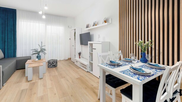A modern living room with a sofa, a dining table set for two, a TV, and a wooden wall panel. - Cozy Studio with a Balcony | Parking (Grzybowo)