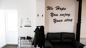 Een kamer met een zwarte leren bank, een wandkapstok met jassen, een schoenenrek met schoenen, een deur en een muur met een motiverende quote.