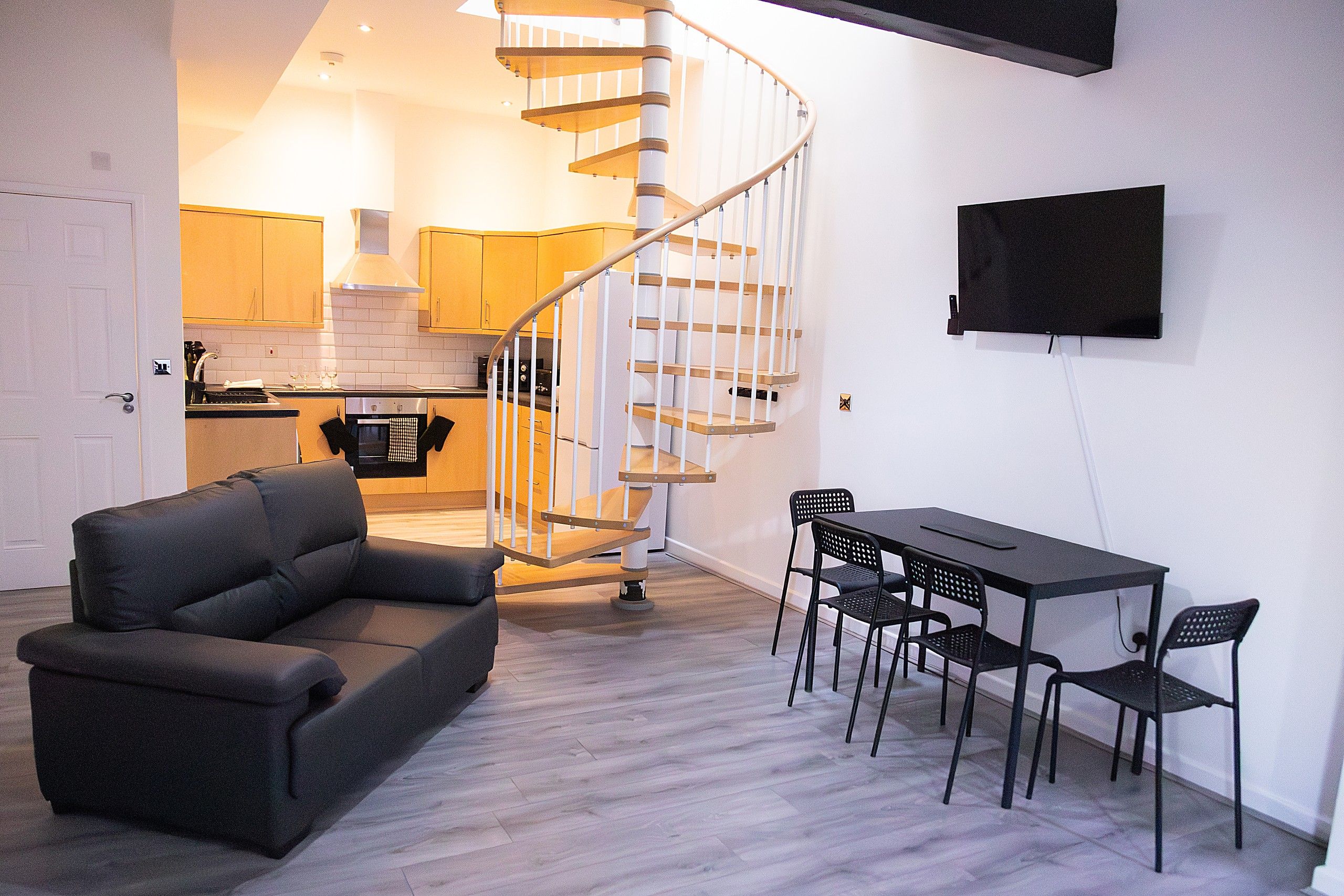 A modern living space with a spiral staircase, a dining area with a table and chairs, and a kitchen in the background.